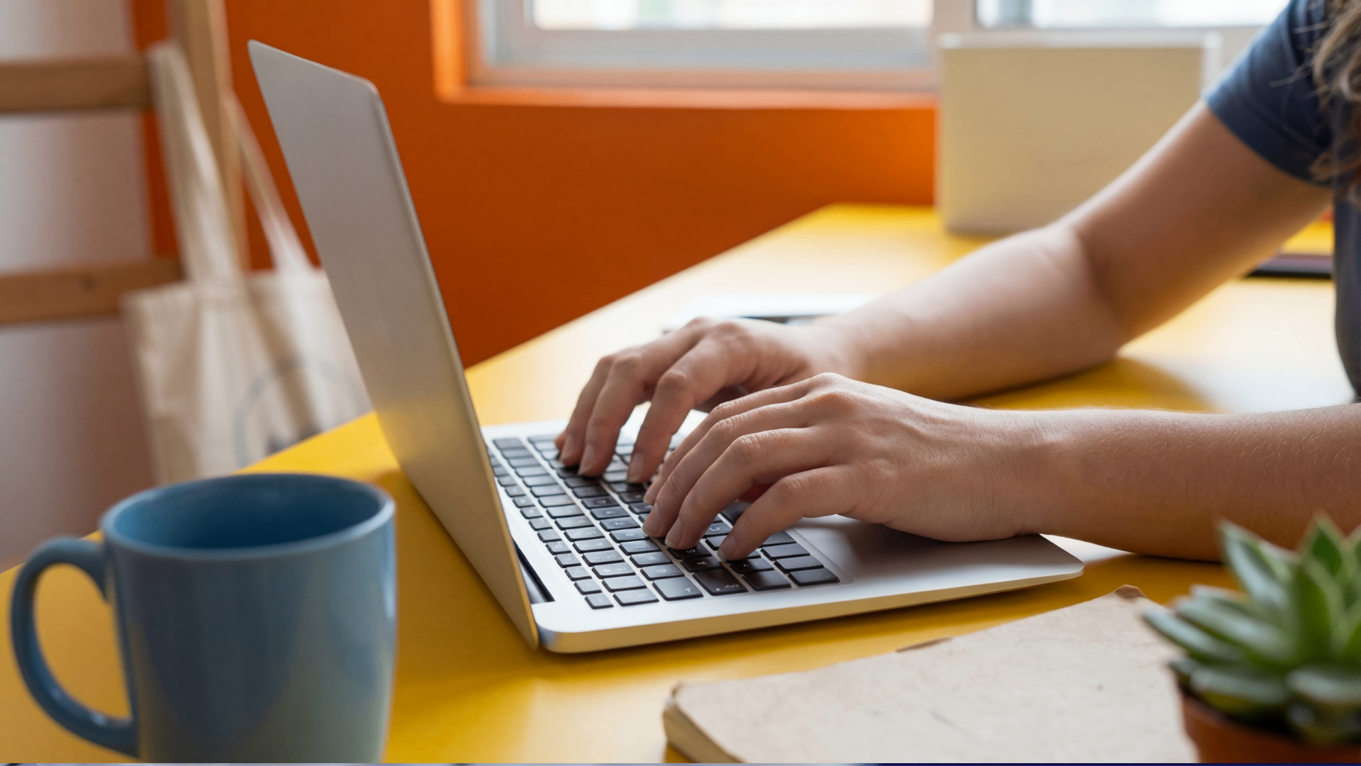 Side profile of hands typing on a laptop on a yellow desk next to a blue mug
