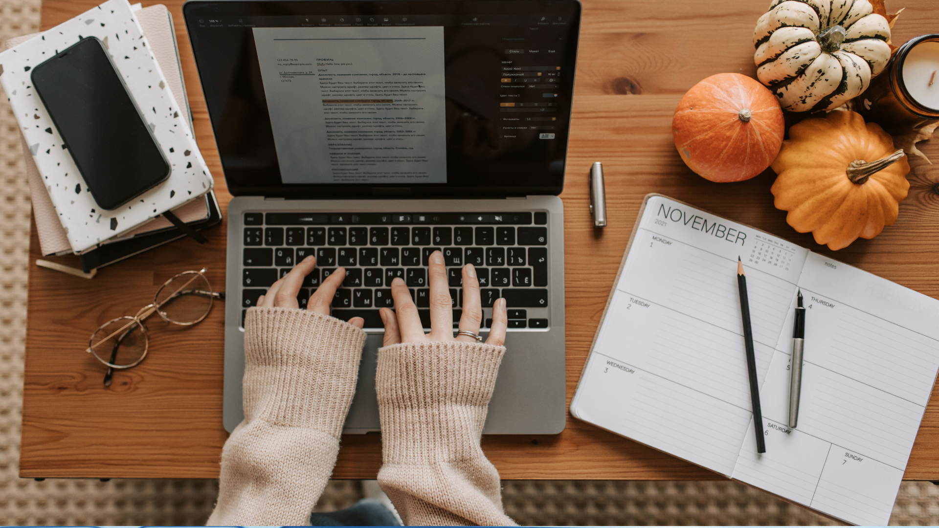 Top-down view of hands in beige sweater typing on a laptop surrounded by a calendar, glasses, and decorative pumpkins